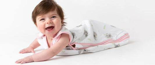 Un neonato sorridente disteso a pancia in giù, indossando un sacco nanna bianco con motivi di animali, mentre esplora e gioca.