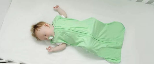 Un neonato dorme tranquillamente in una culla, indossando un sacco nanna verde e con un ciuccio in bocca, trasmettendo una sensazione di serenità e comfort.