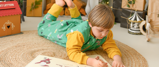 Un bambino, indossando un sacco nanna verde con motivi invernali, è sdraiato a terra disegnando su un libro, circondato da decorazioni natalizie, in un'atmosfera festiva.