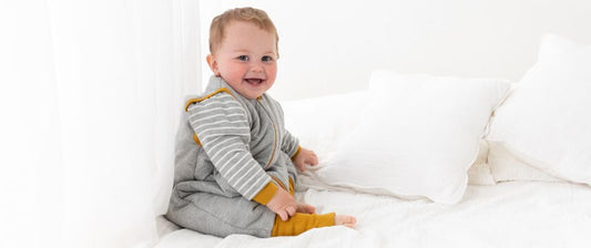Un bambino sorride seduto su un letto, indossando un sacco nanna grigio con dettagli gialli, in un ambiente luminoso e accogliente.