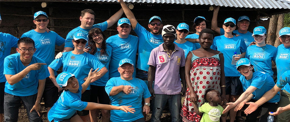 Un gruppo di persone in magliette blu con la scritta "Business for Good" posa sorridendo con una famiglia locale in Kenya, in un ambiente di collaborazione.