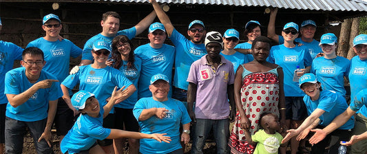 Un gruppo di persone in magliette blu con la scritta "Business for Good" posa sorridendo con una famiglia locale in Kenya, in un ambiente di collaborazione.