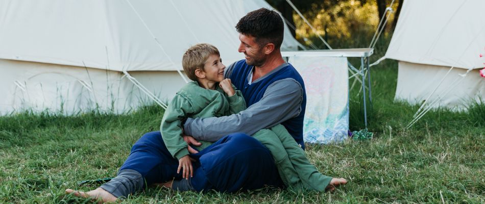 Un padre e suo figlio siedono sull'erba, sorridendo e abbracciandosi, davanti a una tenda da campeggio in un ambiente naturale e tranquillo, creando un momento di affetto.