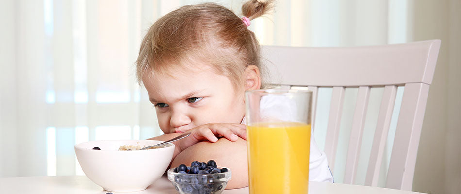 Una bambina guarda con espressione scontenta il suo piatto di cereali, con un bicchiere di succo d'arancia e una ciotola di mirtilli accanto, mostrando disinteresse per il pasto.