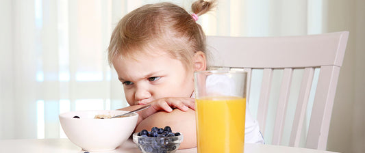 Una bambina guarda con espressione scontenta il suo piatto di cereali, con un bicchiere di succo d'arancia e una ciotola di mirtilli accanto, mostrando disinteresse per il pasto.