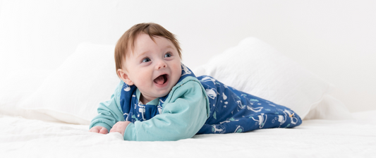Un bambino sorride sdraiato a pancia in giù su un letto, indossando un sacco nanna blu con disegni, creando un'immagine di comfort e felicità in un ambiente luminoso.