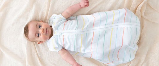 Un neonato sdraiato su una coperta, indossa un sacco nanna bianco con strisce colorate, guardando l'obiettivo con espressione tranquilla e serena.