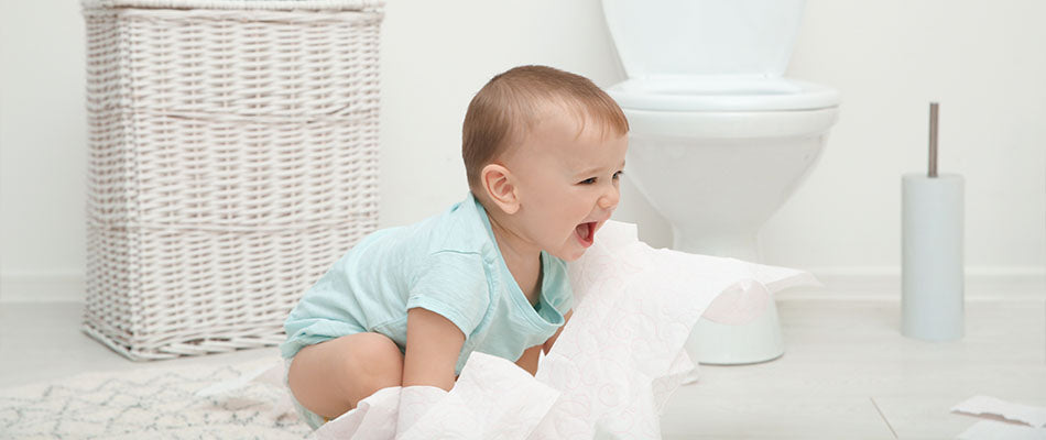 Un bambino sorride seduto sul pavimento del bagno, giocando con carta igienica srotolata, circondato da un'atmosfera divertente e vivace accanto al vasino.