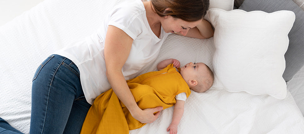 Una madre distesa accanto al suo bambino, che indossa un sacco nanna giallo, mentre si guardano dolcemente su un letto bianco.