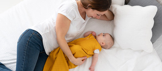 Una madre distesa accanto al suo bambino, che indossa un sacco nanna giallo, mentre si guardano dolcemente su un letto bianco.