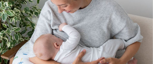 Una madre sorride dolcemente mentre allatta il suo bambino, tenendolo in braccio con un cuscino da allattamento, creando un'atmosfera rilassante e affettuosa.