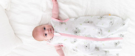 Un neonato è sdraiato su un letto bianco, indossando un sacco nanna bianco con motivi di koala e bordi rosa, guardando verso l'alto con espressione tranquilla.