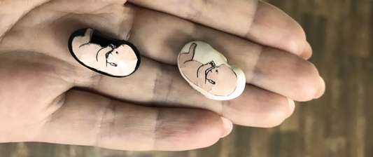 Due mani tengono due piccoli disegni di un embrione umano, rappresentati su adesivi ovali con sfondo bianco e nero.
