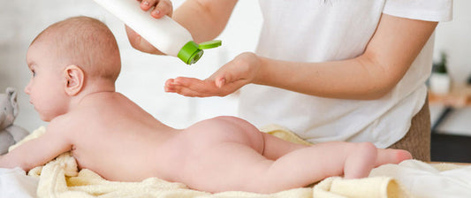 Un neonato è sdraiato a pancia in giù su un asciugamano giallo, mentre un adulto versa crema per il corpo sulla mano, pronto per massaggiare delicatamente la pelle del bambino.