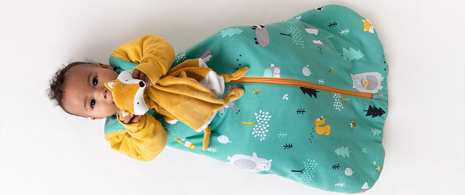 Un bambino sdraiato indossa un sacco nanna verde con disegni di animali della foresta e tiene in mano un peluche, guardando verso l'alto con un'espressione serena.