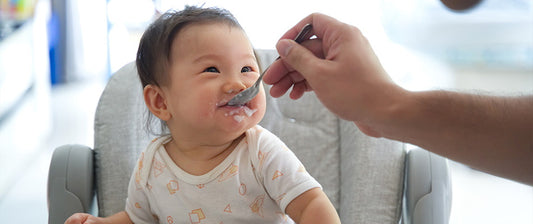 Un bambino sorride mentre viene nutrito con un cucchiaio da un adulto, seduto su un seggiolone, con un po' di cibo sul viso che aggiunge un tocco divertente alla scena.