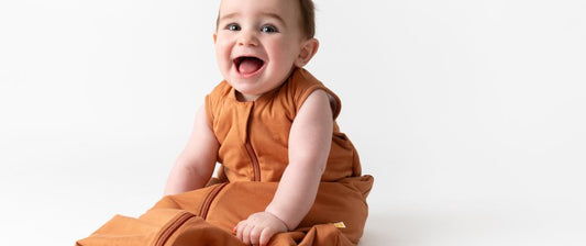 Un bambino sorride seduto, indossando un sacco nanna arancione su sfondo bianco, esprimendo gioia e comfort, con un ambiente semplice e luminoso.