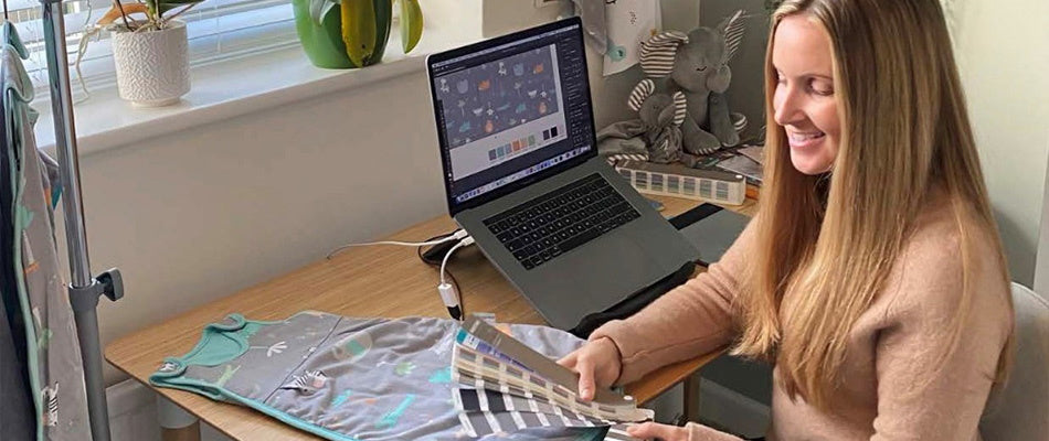 Una designer sorride mentre lavora al computer, scegliendo campioni di colore per un sacco nanna con un design per bambini, mostrando il processo creativo.