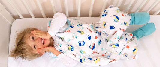 Un bambino sorridente sdraiato in un lettino, indossando un sacco nanna bianco con disegni colorati, mentre gioca felicemente.