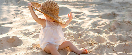 Un bambino seduto sulla sabbia indossa un grande cappello di paglia, cercando di sistemarlo sulla testa, mentre gioca sotto il sole in una giornata estiva in spiaggia.
