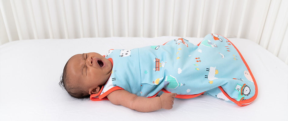 Un neonato che sbadiglia mentre è disteso nel suo lettino, indossando un sacco nanna azzurro con motivi colorati, pronto per dormire.
