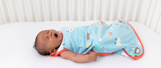 Un neonato che sbadiglia mentre è disteso nel suo lettino, indossando un sacco nanna azzurro con motivi colorati, pronto per dormire.