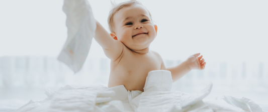 Un bambino sorridente gioca con dei pannolini, seduto in mezzo a loro, sollevandone uno in aria con espressione felice e vivace.