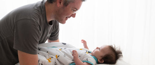  Un papà guarda dolcemente il suo bambino che indossa un sacco nanna bianco con disegni di animali, disteso su un letto bianco e rilassato.