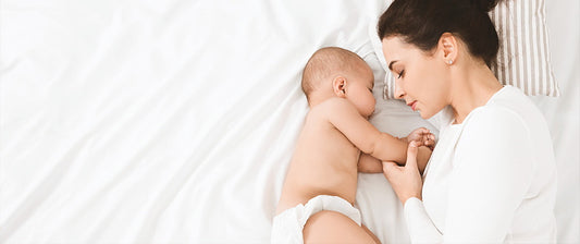 Una madre dorme accanto al suo bambino in un letto bianco, tenendogli dolcemente la mano, creando un'immagine serena e affettuosa di riposo condiviso.