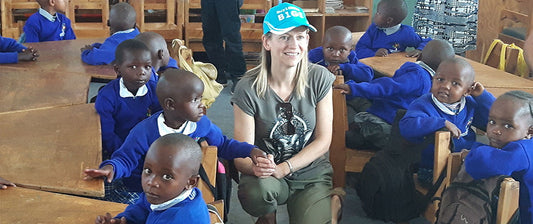 Una donna con un cappello azzurro siede tra un gruppo di bambini vestiti con uniformi blu, sorridendo in una classe, creando un'atmosfera di condivisione e apprendimento.